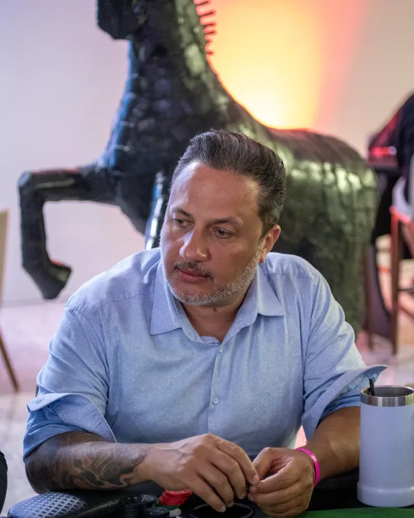 Alex Brito de camisa azul clara, sentado à mesa de poker, com uma escultura de cavalo preto ao fundo